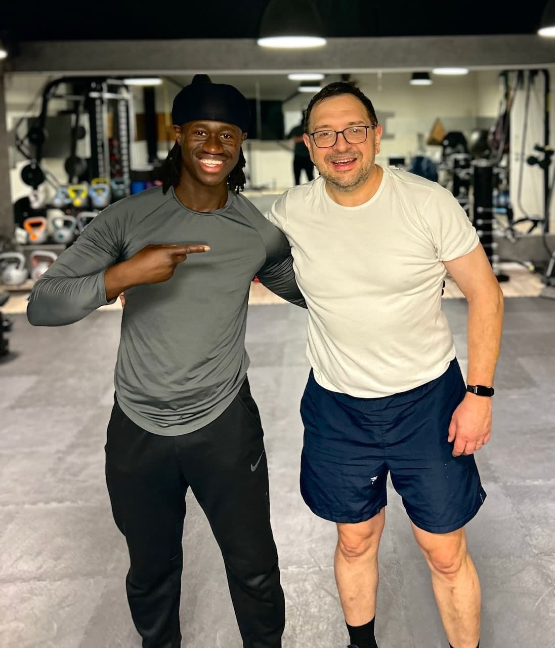 Deux hommes souriants posent ensemble dans une salle de sport, entourés d'équipements d'entraînement.