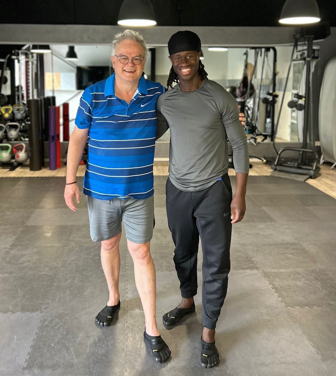 Deux hommes souriants posant dans une salle de sport, portant des vêtements de sport et des chaussures minimalistes.
